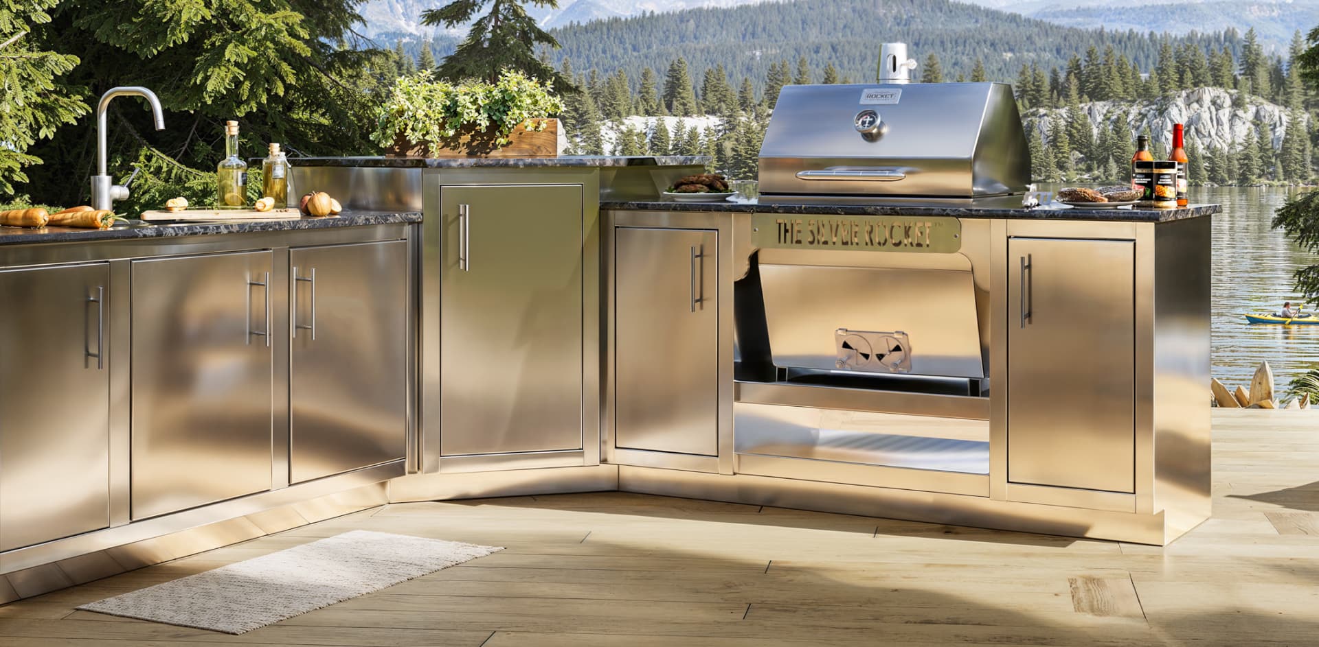Silver Rocket stainless steel grill built into a Rocket Outdoors outdoor kitchen with cabinets, sink, countertop prep space, and a lake view.